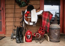 Portrait Of A Black Great Dane Resting On A Farmhouse Porch Beside A Saddle Rack With English Tack Including Saddle, Bridle, Riding Boots Decorated For Christmas- Bells, Lantern And Plaid Blanket