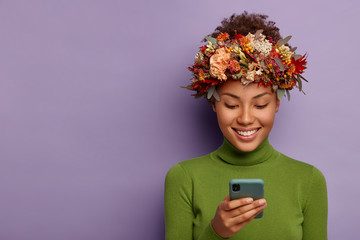 Portrait of glad autumn model wears decorative autumnal wreath, focused in smartphone device, reads good news online, has happy face expression, models over purple studio wall. Technology, season