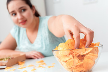 Wall Mural - breaking diet. chubby girl sitting at kitchen table eating chips excited close-up blurred background