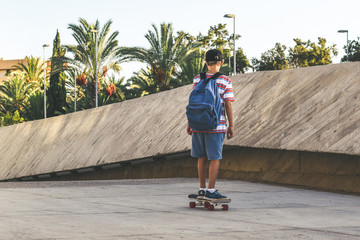 Young boy riding a skate out of school in a urban context. Teen with backpack and rapper hat, having fun with skateboard in a modern city.   Happy student backing home from the institute Youth concept