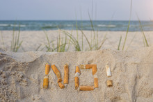 In The Foreground, The Word "no!" Written In The  Sand With Cigarette Stubs,  The Sea Is In The Background