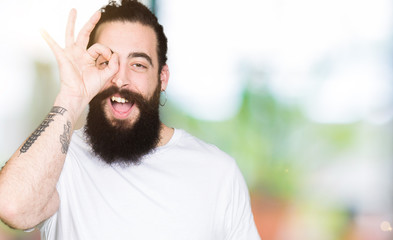 Sticker - Young hipster man with long hair and beard wearing casual white t-shirt doing ok gesture with hand smiling, eye looking through fingers with happy face.