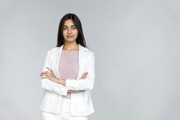 Wall Mural - Confident and successful young adult indian businesswoman in white suit crossed arms on chest, standing isolated on grey background with copy space and looking at camera