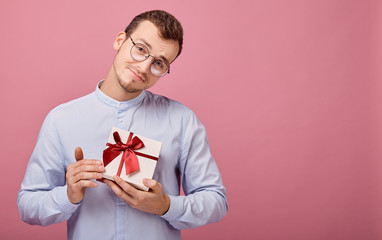 A surprised man in shirt with glasses holds a gift in a box with lid and red bow in his hands, looking on present. Holiday, rejoicing. Smiling, happiness. Give a gift. Guy bowed his head.