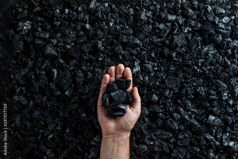 Male hand with coal on the background of a heap of coal, coal mining in an open pit quarry, copy space. Fossil fuels, environmental pollution. - obrazy, fototapety, plakaty 