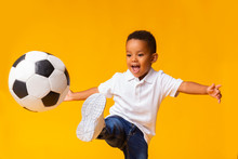 Adorable Little Boy Playing Football, Hitting Ball Over Yellow Background