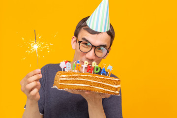 Wall Mural - Portrait of a funny positive guy with a paper cap and glasses holding a congratulatory homemade cake in his hands on a yellow background. Concept and fun and celebration.