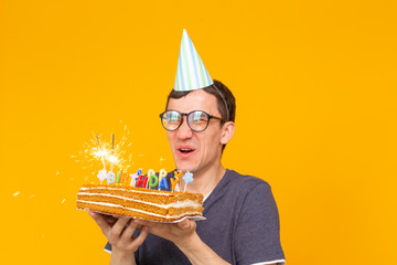 Wall Mural - Positive funny young asian guy with a cap and a burning candle and a homemade cake in his hands posing on a yellow background. Anniversary and birthday concept.