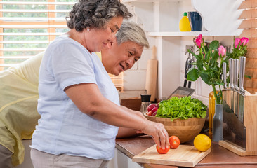 Asian senior wife slicing tomatoes on wooden cutting  board and using tablet computer to searching menu recipe.senior with technology lfiestyle.aging at home