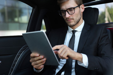 Thoughtful young businessman sitting in the luxe car and using his tablet.