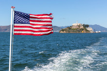 The Stars And Stripes Flag Flying From The Back Of A Boat With Alcatraz Island Out Of Focus Behind.