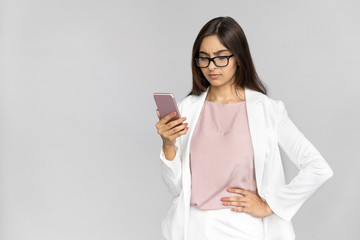 Confused serious angry young adult indian businesswoman user customer in formal wear suit stand isolated on grey background with copy space holding smartphone in hand looking at screen feel annoyed