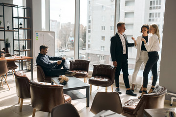 Business people meeting, talking while standing in front of the panorama window, while handsome cheerful man sitting on the armchair and using his tablet. copy space.conversation concept