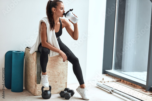 Fotografía Young woman in leggins with towel on shoulders drinking water after fitness trai