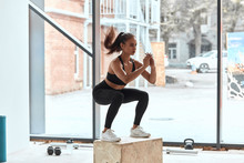 Jumping On Fit Box In Process. Young Athletic Woman Do Squats On Wooden Box In Gym. Window Background