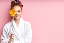 Young Smiling Woman Holding Daisy Yellow Flower Isolated Over Pink Background. Female Wearing White Bathrobe, Curlers On Hair After Shower. Natural Beauty