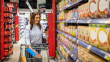 Pretty Young Woman Buying Groceries In A Supermarket/mall/grocery Store. Shopping List. Smiling Beautiful Woman Stands With Phone Near Products In Store