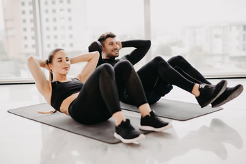 Wall Mural - Young couple make sits up at gym. Attractive woman and handsome muscular man are training in light modern gym. Abs exercises.