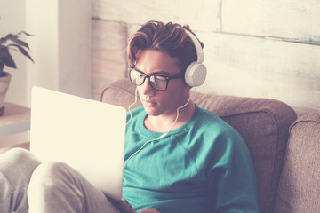 Young student at home with modern laptop computer and headphone studying with internet cnnection - handsome boy sitting on the sofa with technology indoor