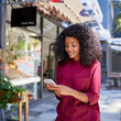 © marvent - Smiling young woman standing on a sidewalk reading text messages