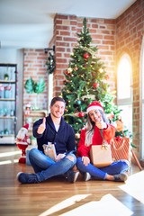 Poster - Young couple wearing santa claus hat sitting on the floor around christmas tree at home doing happy thumbs up gesture with hand. Approving expression looking at the camera with showing success.