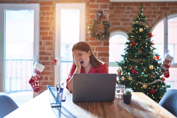 Poster - Beautiful woman sitting at the table working with laptop at home around christmas tree Yawning tired covering half face, eye and mouth with hand. Face hurts in pain.