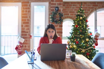 Poster - Beautiful woman sitting at the table working with laptop at home around christmas tree In shock face, looking skeptical and sarcastic, surprised with open mouth