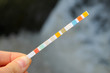 Strip for water quality measurement held in fingers (female hand). Ten multicolor squares for chemical testing of different reagents in water to identify its purity. Background of waterfall.