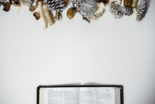 Overhead Shot Of An Open Bible On A White Surface With Pine Cones And An Ornament On The Top