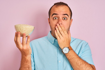 Sticker - Young man holding bowl with rice standing over isolated pink background cover mouth with hand shocked with shame for mistake, expression of fear, scared in silence, secret concept