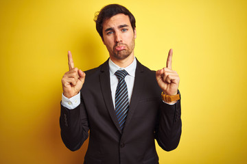 Sticker - Young handsome businessman wearing suit and tie standing over isolated yellow background Pointing up looking sad and upset, indicating direction with fingers, unhappy and depressed.