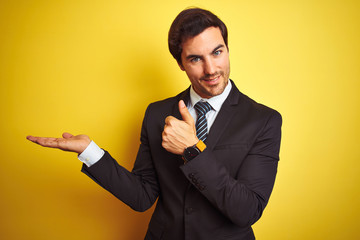 Wall Mural - Young handsome businessman wearing suit and tie standing over isolated yellow background Showing palm hand and doing ok gesture with thumbs up, smiling happy and cheerful