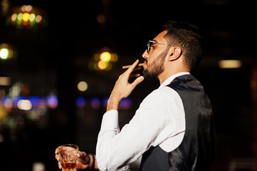 Wall Mural - Handsome well-dressed arabian man with glass of whiskey and cigar posed at pub.