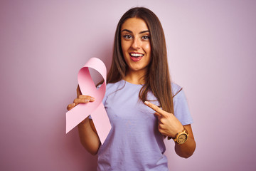 Wall Mural - Young beautiful woman holding cancer ribbon standing over isolated pink background very happy pointing with hand and finger