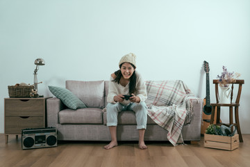 Wall Mural - copyspace woman winter relax indoor. cheerful young beautiful asian chinese girl in warm knitted clothes at home living room on couch. excited lady in beanie playing video game and keeping mouth open