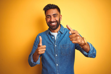 Wall Mural - Young indian man wearing denim shirt standing over isolated yellow background pointing fingers to camera with happy and funny face. Good energy and vibes.