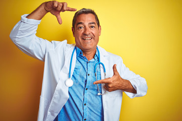 Wall Mural - Handsome middle age doctor man wearing stethoscope over isolated yellow background smiling making frame with hands and fingers with happy face. Creativity and photography concept.