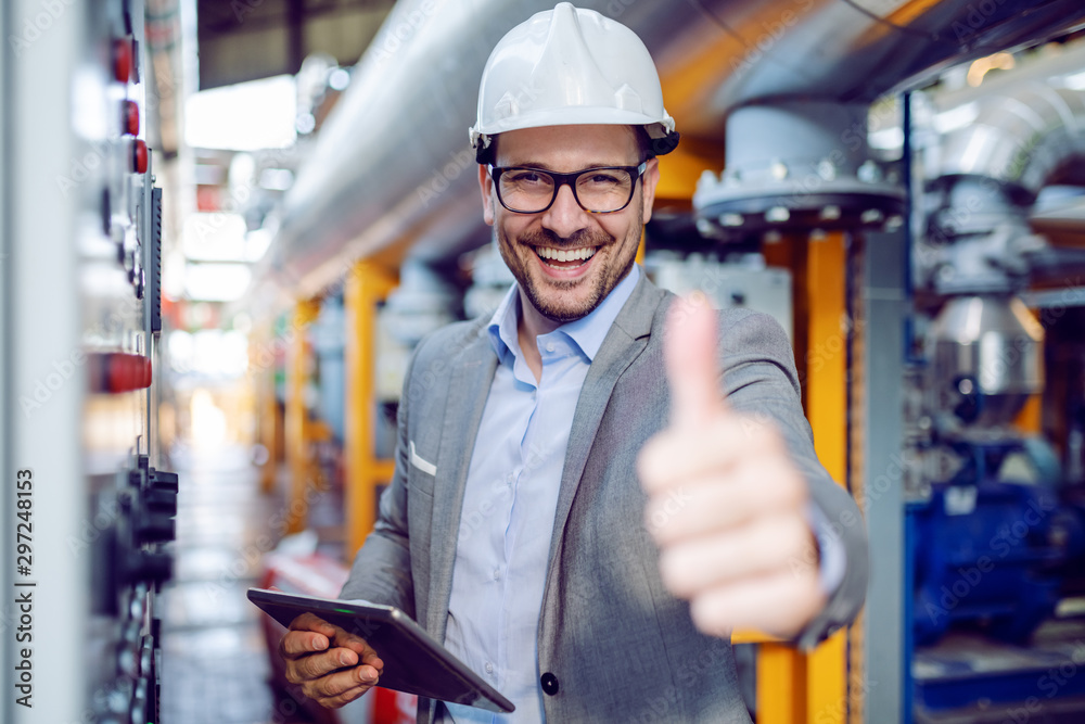 Successful caucasian supervisor in suit and with helmet holding tablet and showing thumbs up while standing next to dashboard in power plant. - obrazy, fototapety, plakaty 