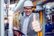 Successful caucasian supervisor in suit and with helmet holding tablet and showing thumbs up while standing next to dashboard in power plant.