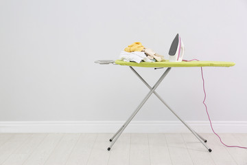 Board with modern iron and pile of clothes near light wall, space for text. Laundry day