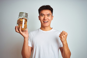 Young asian chinese man holding jar of cookies standing over isolated white background screaming proud and celebrating victory and success very excited, cheering emotion