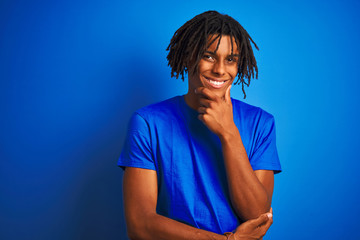 Wall Mural - Afro american man with dreadlocks wearing t-shirt standing over isolated blue background looking confident at the camera smiling with crossed arms and hand raised on chin. Thinking positive.