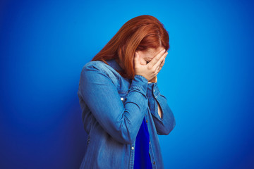 Canvas Print - Young beautiful redhead woman wearing denim shirt standing over blue isolated background with sad expression covering face with hands while crying. Depression concept.