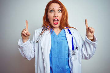 Wall Mural - Young redhead doctor woman using stethoscope over white isolated background smiling amazed and surprised and pointing up with fingers and raised arms.