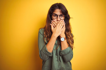 Wall Mural - Young beautiful woman wearing green shirt and glasses over yelllow isolated background shocked covering mouth with hands for mistake. Secret concept.