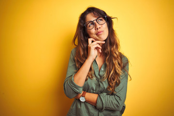 Wall Mural - Young beautiful woman wearing green shirt and glasses over yelllow isolated background with hand on chin thinking about question, pensive expression. Smiling with thoughtful face. Doubt concept.