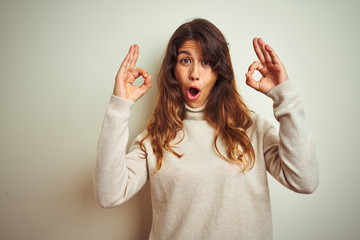 Wall Mural - Young beautiful woman wearing winter sweater standing over white isolated background looking surprised and shocked doing ok approval symbol with fingers. Crazy expression