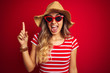 © Krakenimages.com - Young beautiful woman wearing sunglasses and summer hat over red isolated background pointing finger up with successful idea. Exited and happy. Number one.