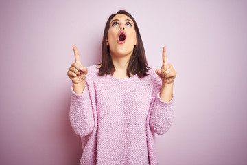 Sticker - Young beautiful brunette woman wearing a sweater over pink isolated background amazed and surprised looking up and pointing with fingers and raised arms.