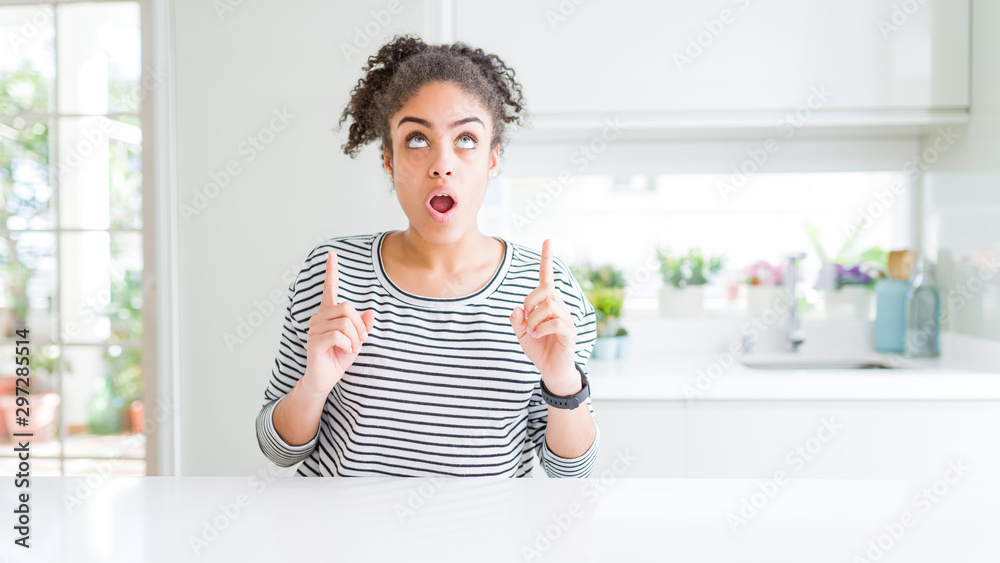 Beautiful african american woman with afro hair wearing casual striped sweater amazed and surprised looking up and pointing with fingers and raised arms.